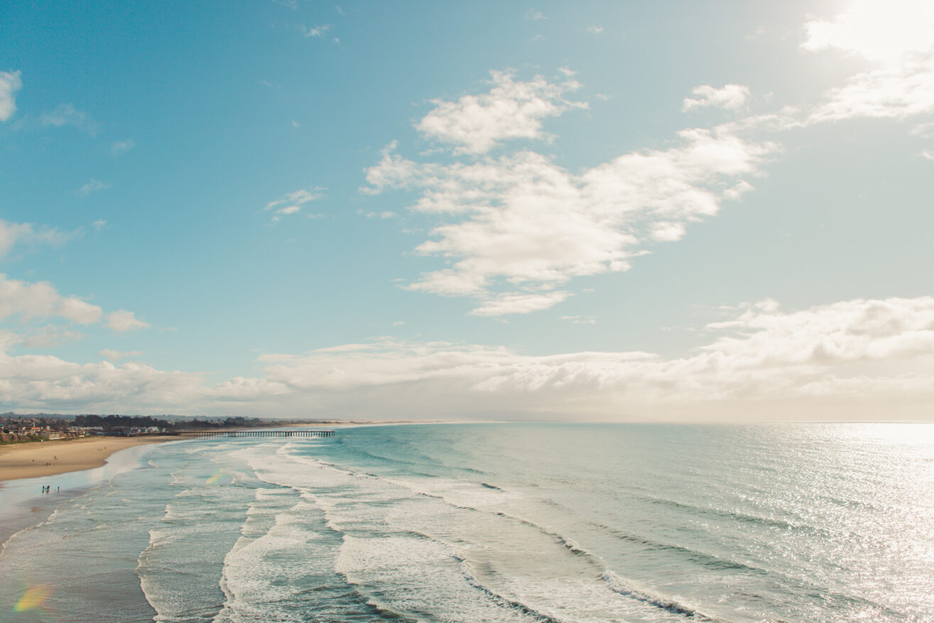 Wide sandy beach with gentle waves rolling onto the shore under a bright blue sky with scattered clouds. Small groups of people walk along the shoreline, and sunlight sparkles on the water.