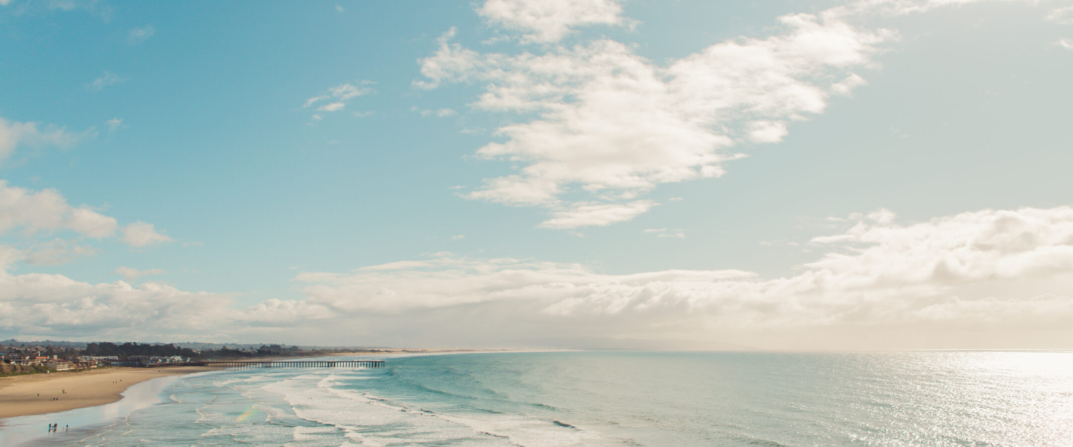 Wide sandy beach with gentle waves rolling onto the shore under a bright blue sky with scattered clouds. Small groups of people walk along the shoreline, and sunlight sparkles on the water.
