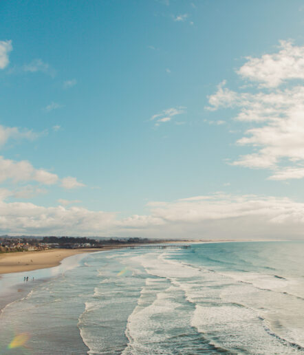 Wide sandy beach with gentle waves rolling onto shore, a long pier stretches into the water. A few people walk along the shoreline under a blue sky with scattered clouds. Houses and trees are visible in the distance.
