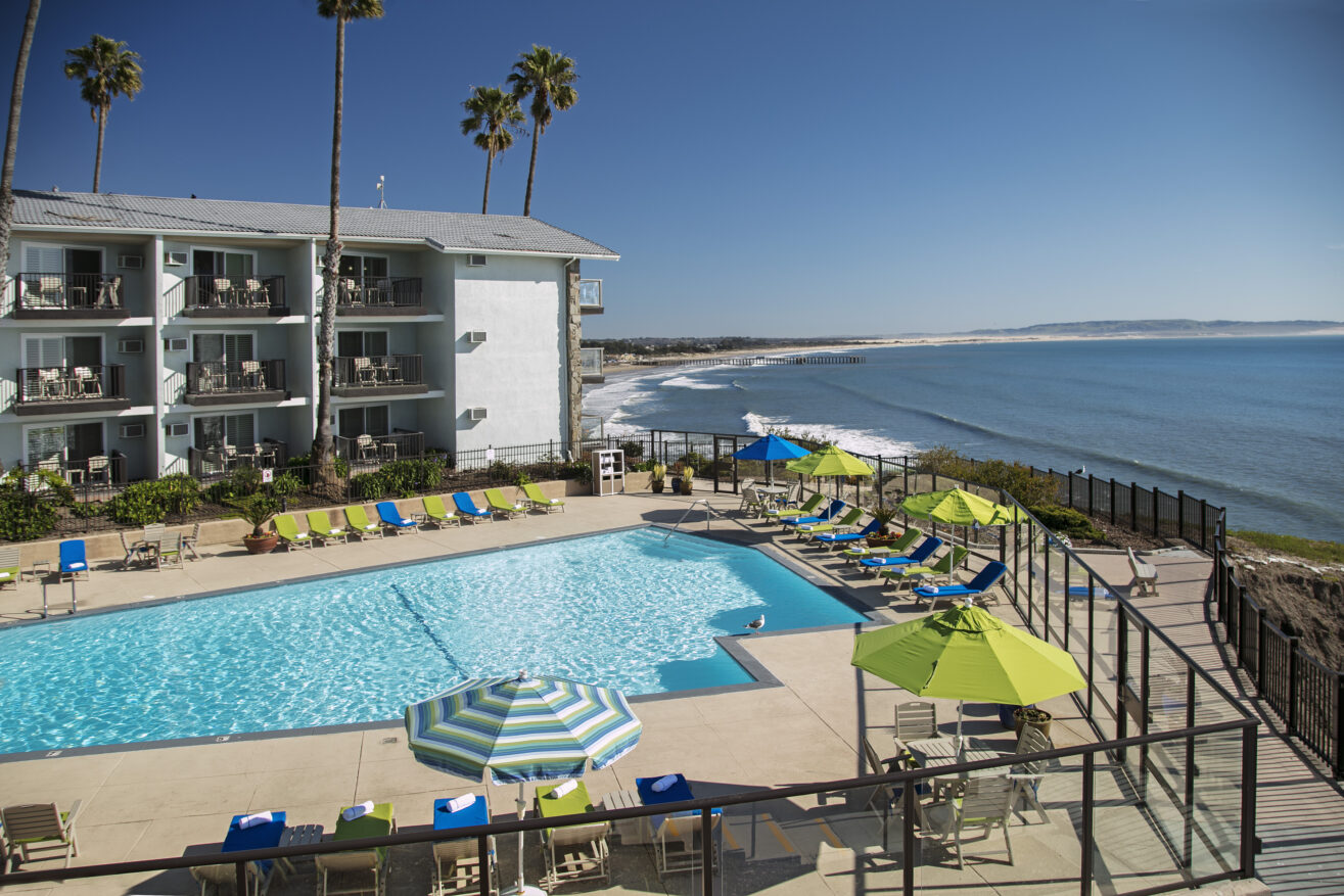 A large outdoor pool with colorful lounge chairs and umbrellas overlooks the ocean near a beachfront hotel under a clear blue sky, with waves rolling onto the sandy shore in the background.