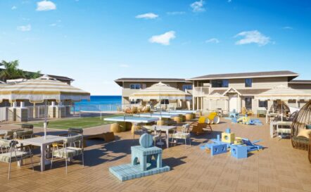 A spacious outdoor patio with tables, chairs, sun umbrellas, and childrens play equipment, overlooking a pool and a two-story building with the ocean visible in the background under a clear blue sky.