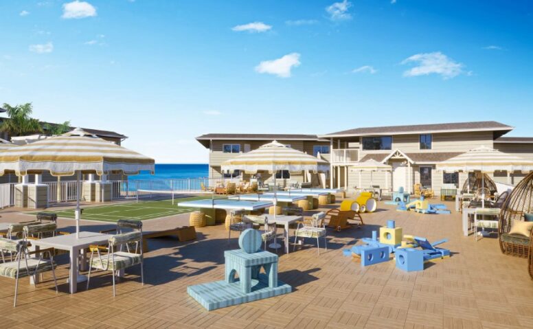 A spacious outdoor patio with tables, chairs, sun umbrellas, and childrens play equipment, overlooking a pool and a two-story building with the ocean visible in the background under a clear blue sky.