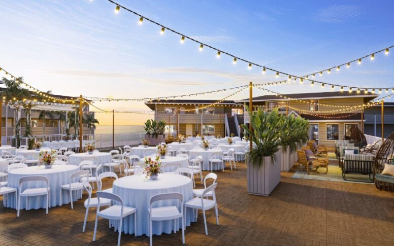 An outdoor event space with round tables covered in white cloths and surrounded by white chairs, decorated with floral centerpieces. String lights are hung above, and the sky is clear at sunset.