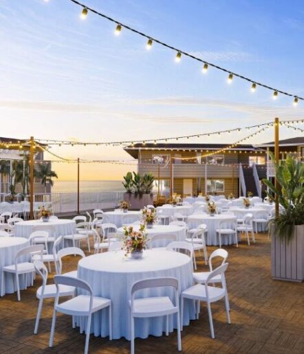 Outdoor event space at sunset with round tables covered in white cloths, white chairs, flower centerpieces, string lights overhead, and a view of modern buildings and the ocean in the background.