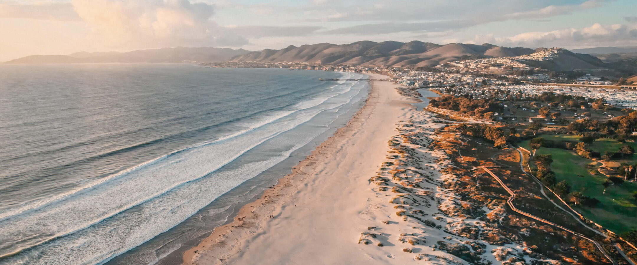 Aerial view of a wide sandy beach with gentle waves meeting the shore, a coastal town, rolling hills, and scattered trees under a partly cloudy sky at sunset.