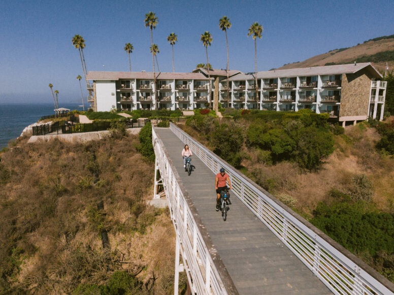 Two people ride bikes on a wooden bridge over a coastal cliff, with a multi-story seaside hotel, palm trees, and the ocean visible in the background under a clear blue sky.
