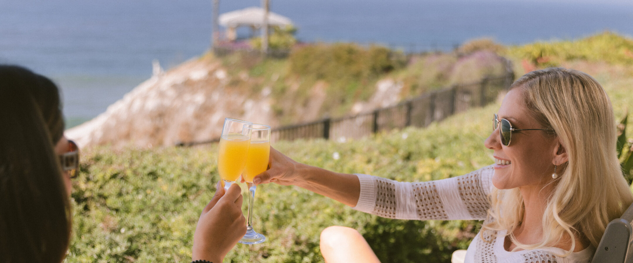 Two women sit outdoors by the ocean, wearing sunglasses and light clothing, smiling and clinking glasses of orange juice against a scenic backdrop of greenery, cliffs, and blue skies.