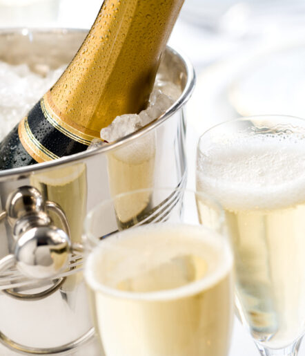 A close-up of a champagne bottle in an ice bucket next to two filled glasses of champagne, with bubbles visible in the drinks. The setting appears elegant and festive.