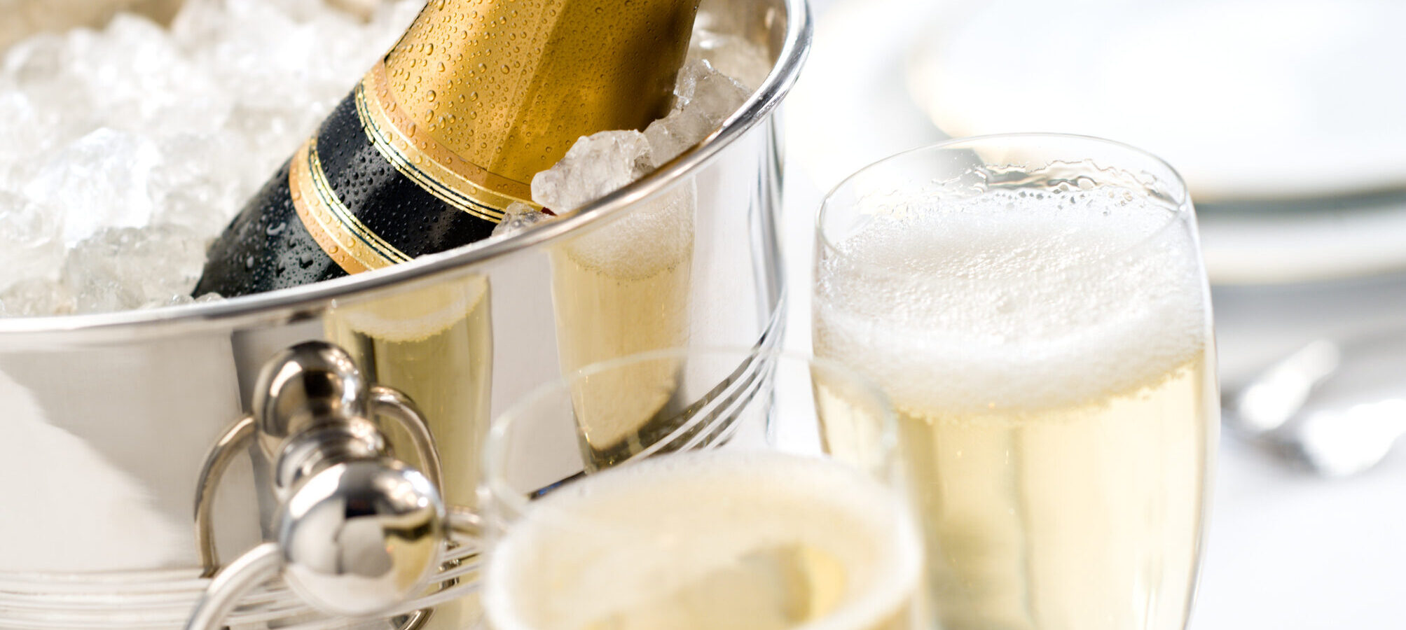 A bottle of champagne sits in an ice-filled silver bucket next to two filled champagne flutes, with plates and cutlery blurred in the background.