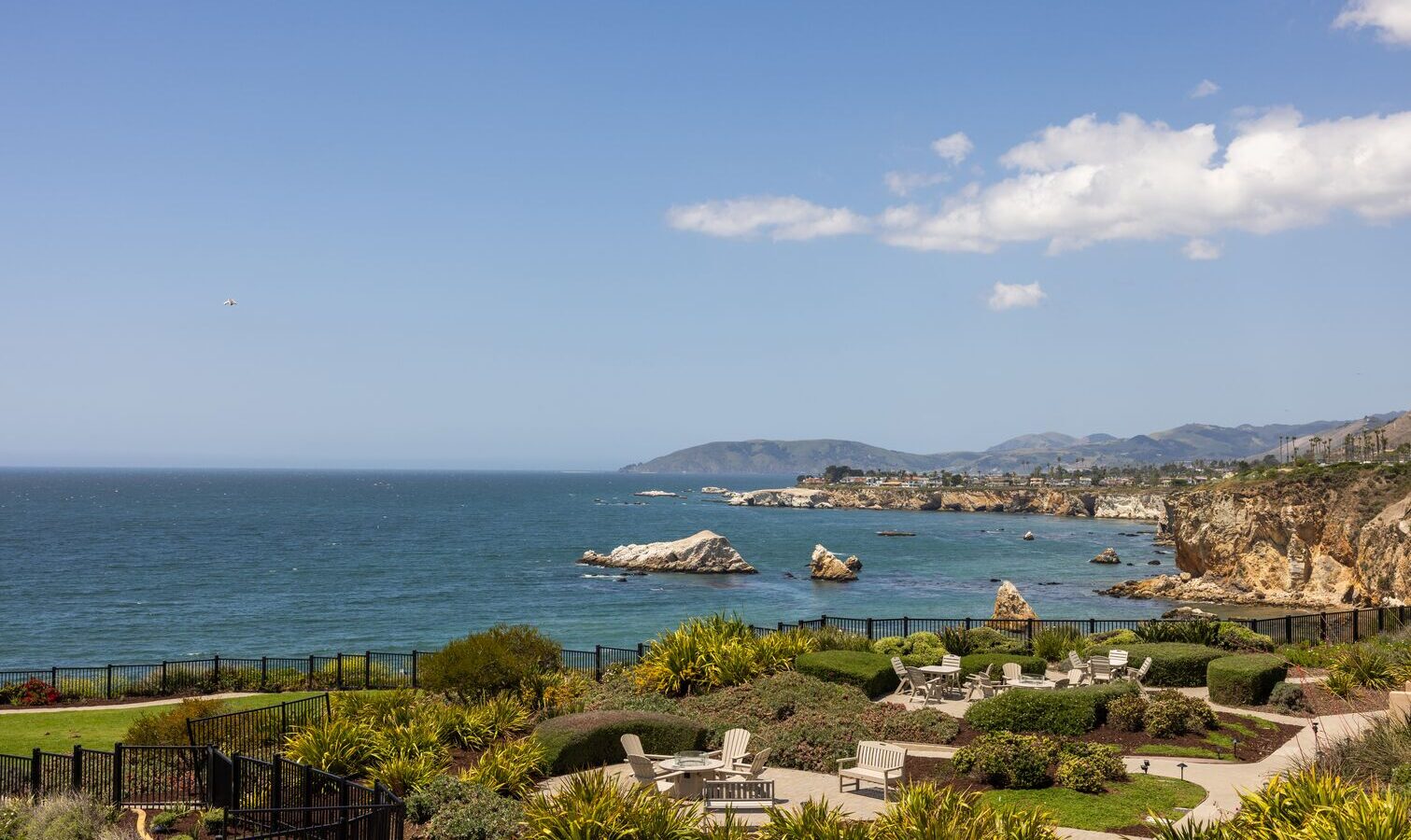 A coastal view with landscaped gardens, patio seating, and fences in the foreground overlooking the ocean, rocky shoreline, and distant hills under a blue sky with scattered clouds.