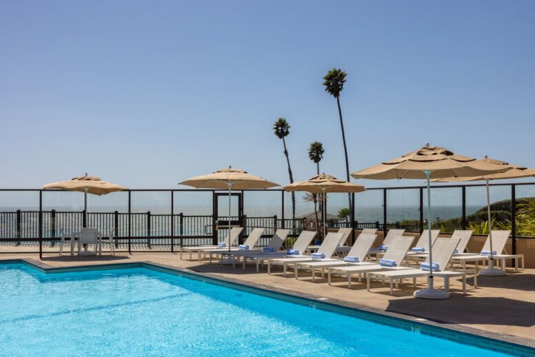 A row of white lounge chairs with rolled towels sits beside a clear blue swimming pool, shaded by beige umbrellas. Palm trees and an ocean view are visible beyond a black fence under a clear sky.