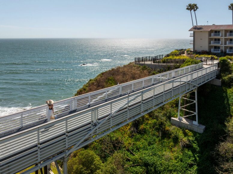 A metal walkway on stilts leads over green shrubs toward a seaside cliff, with an ocean view and a building with balconies on the right under a clear blue sky.