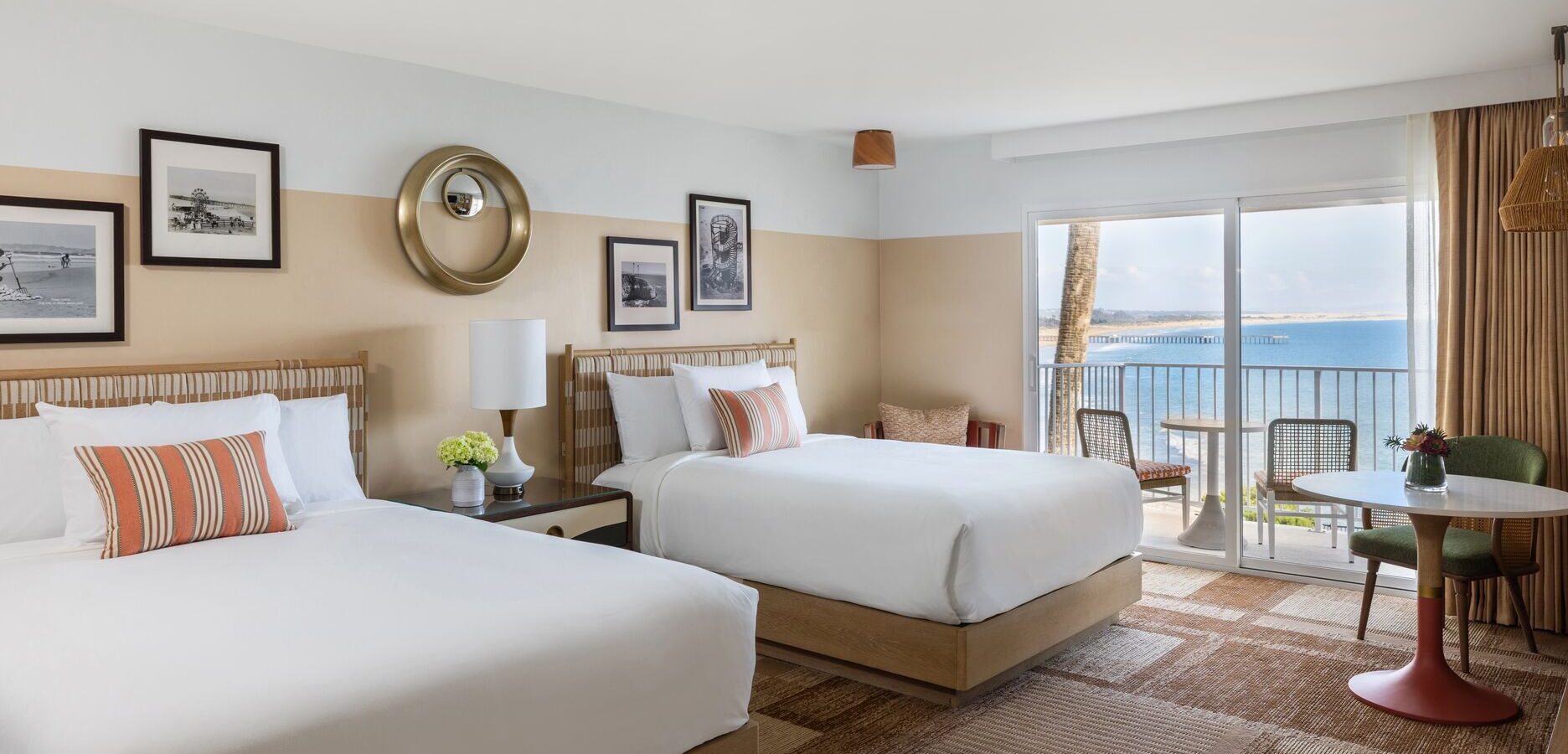 Bright hotel room with two double beds, striped accent pillows, framed art on the walls, a lamp, a small round table with chairs, and a balcony overlooking the ocean under a clear sky.