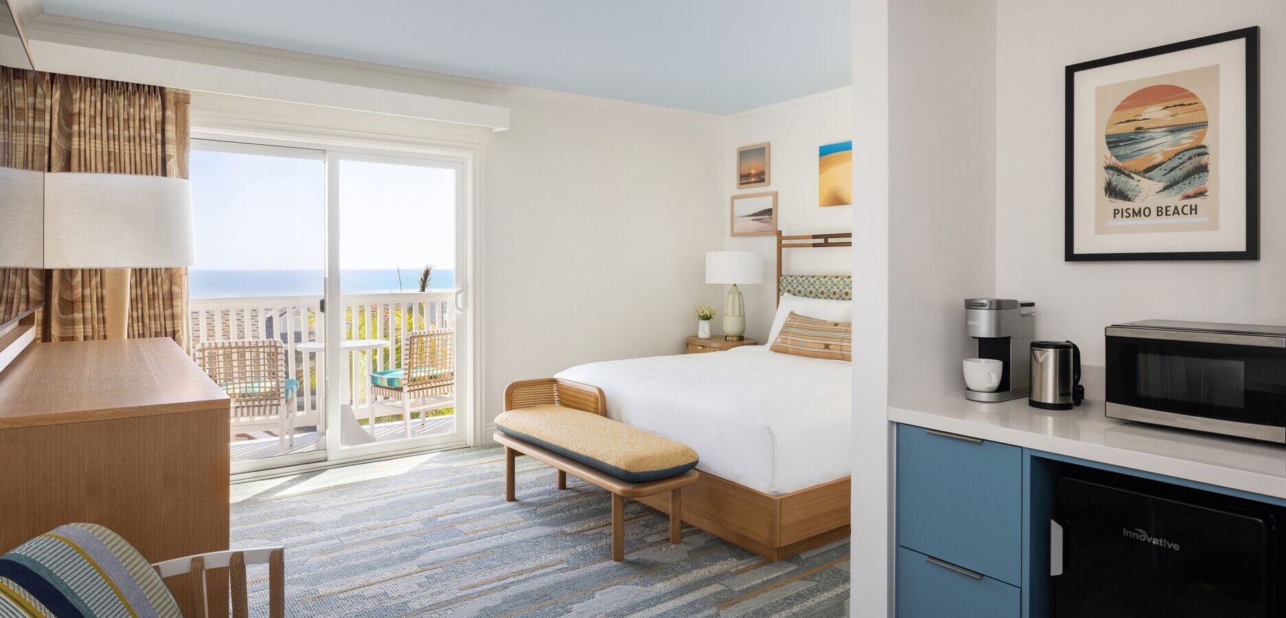A bright, modern hotel room with a king bed, blue accents, a small table and chairs, a microwave, coffee maker, mini fridge, patio doors, and a balcony with an ocean view. Beach-themed artwork decorates the walls.
