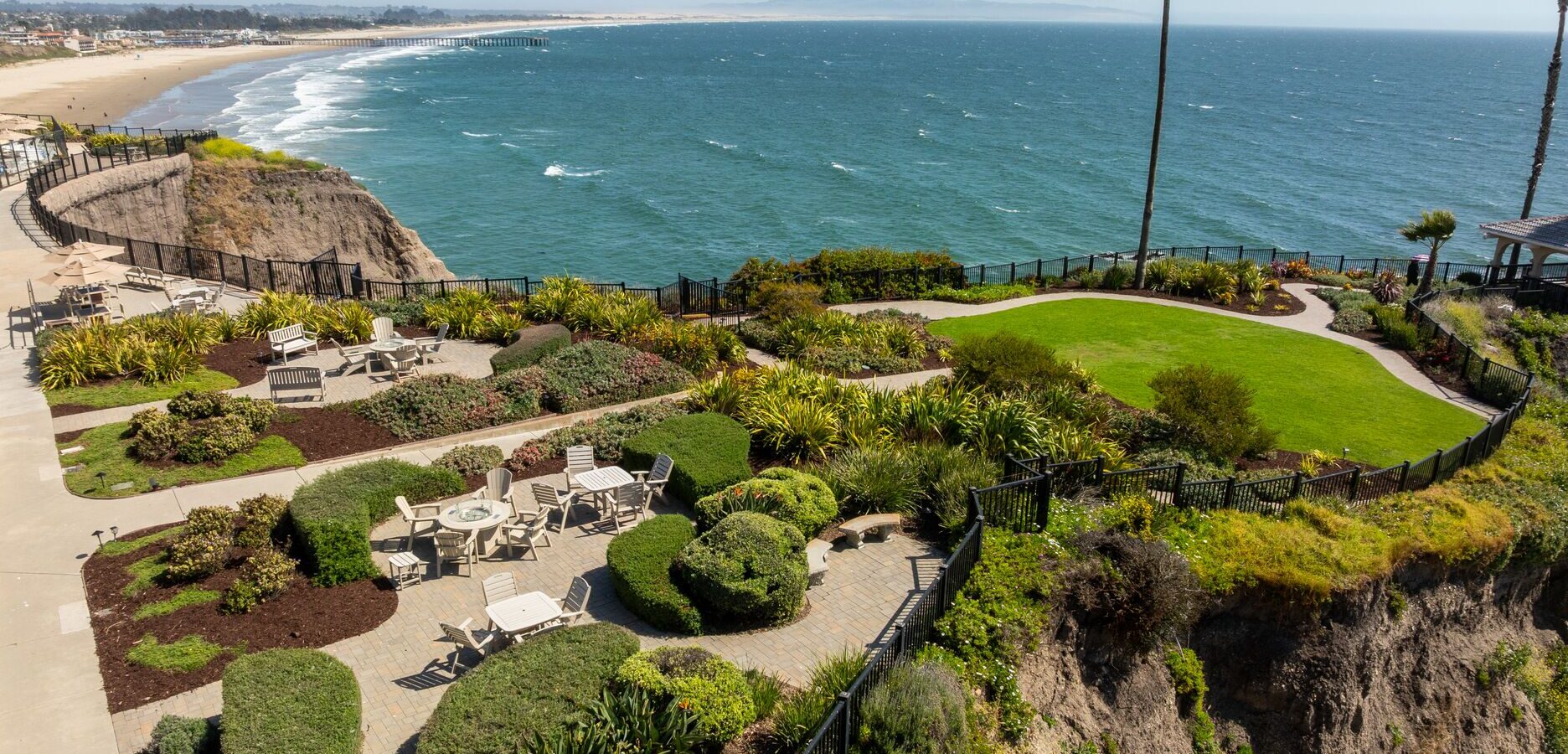 A landscaped garden with patios and seating overlooks the ocean from a cliff, with a sandy beach and distant horizon visible under a clear blue sky.