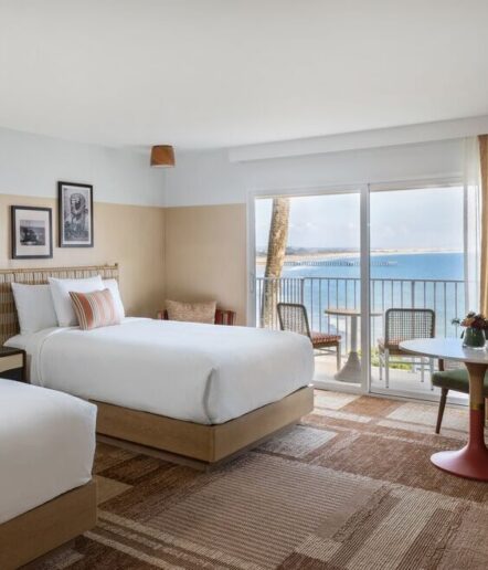 A bright hotel room with two double beds, modern decor, framed photos on the wall, and a sliding glass door opening to a balcony with two chairs and a sea view.