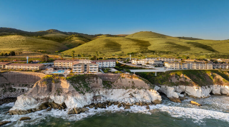 Aerial view of buildings on top of dramatic seaside cliffs, with rolling green hills in the background and waves crashing against the rocky shore below, under a clear blue sky.